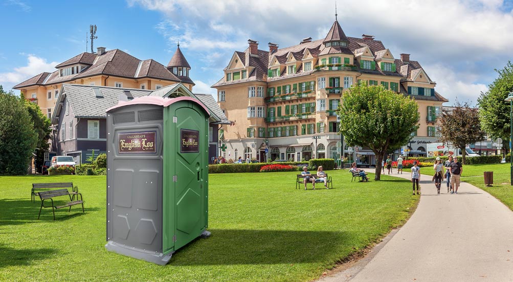 English Loo | English Loo Portable Toilet at a grassy area of a park on a sunny day, with a distinctive building in the background.